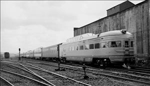 A train car with a rounded back and a glass dome on top. It is at the end of a long line of passenger rail cars, passing a concrete building and rows of empty railroad tracks. 